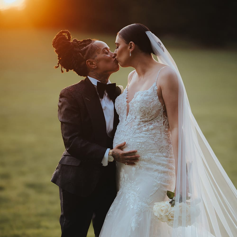Bride in lace sleeveless v neck Enzoani wedding dress with tulle finish and church length veil and bride in black snakeskin print tuxedo with black bowtie and white rose boutonniere holding her baby bump