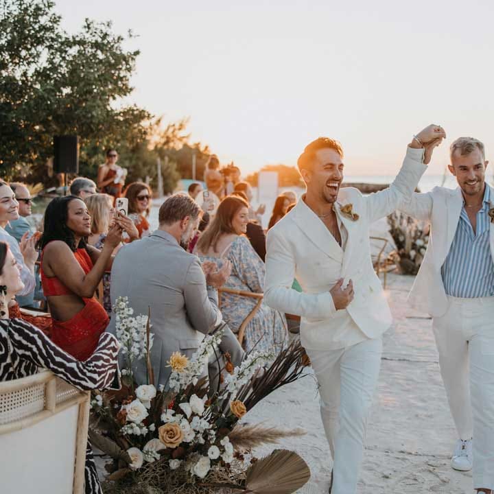 Groom in mens white wedding suit with dusky orange garden rose and dried flower boutonniere, white shirt and silver jewellery holding hands and walking down the aisle with groom in white blazer, light blue and white striped shirt, dusky orange garden rose and dried flower boutonniere and gold jewellery