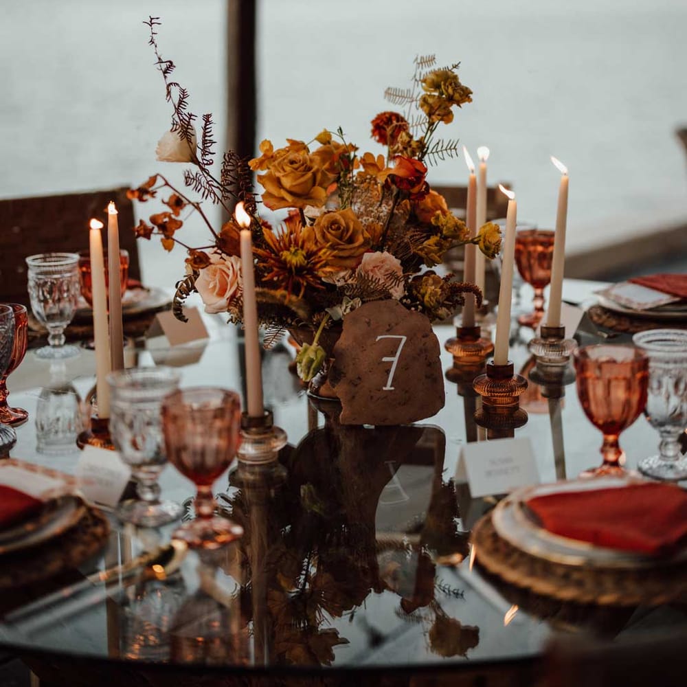 Rustic wedding tablescape with burnt orange dried roses, dried flower and foliage floral centrepieces, wooden slate wedding table numbers, light orange tapered candles and burnt orange wedding napkins 