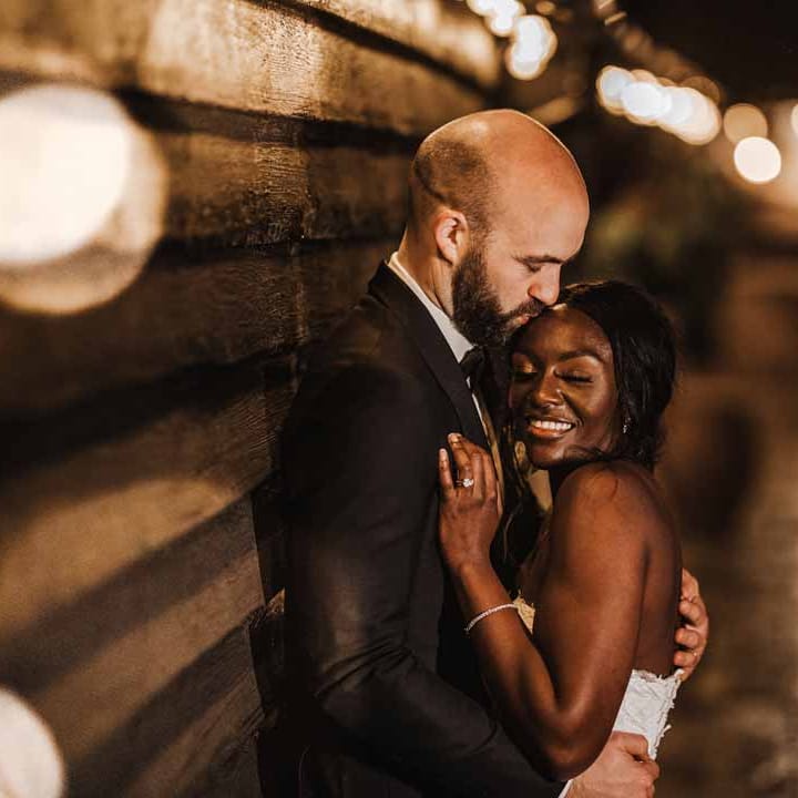 Bride in sweetheart neckline lace wedding dress hugging groom in classic black three piece grooms suit with bow tie and white rose and foliage boutonniere in church at Lains Barn