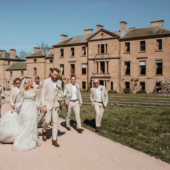 Bride in sage green off shoulder long sleeve wedding dress with groom in light suit with wedding party in sage green wedding outfits walking the grounds in one of the best wedding venues Scotland - Cambo House 
