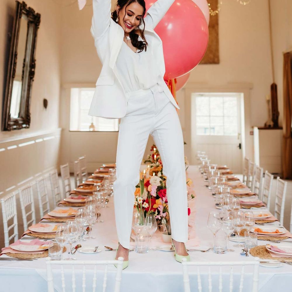 Bride in white bridal suit with closed toe bright green wedding heels standing on wedding tablescape with white wedding tablecloth, burlap place mats, tapered candles, pink and orange wedding stationery, and floral wedding table centrepieces with pink and orange garden roses, orange floribunda, yellow ranunculus flowers, foliage and Iceland poppies 