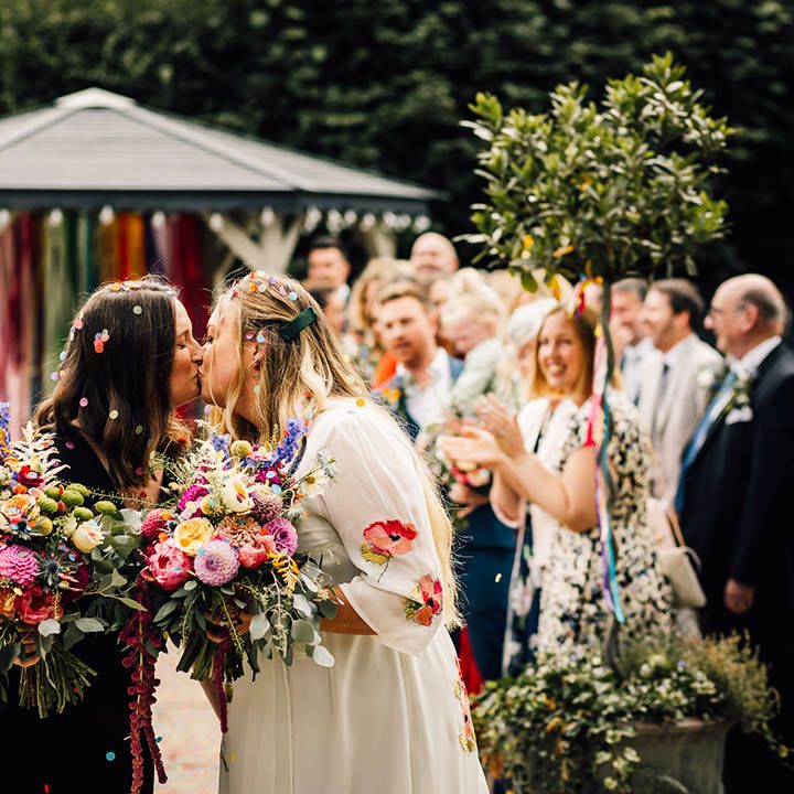 Two brides with one wearing a floral embroidered wedding dress and the other in black bridal jumpsuit share a kiss after their confetti exit 