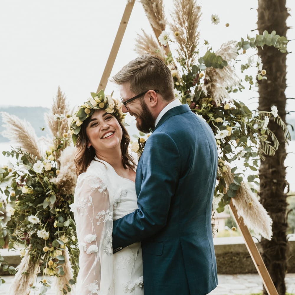 Boho bride in a flower crown and white wedding dress with flowers smiling as her groom embraces her at the altar 