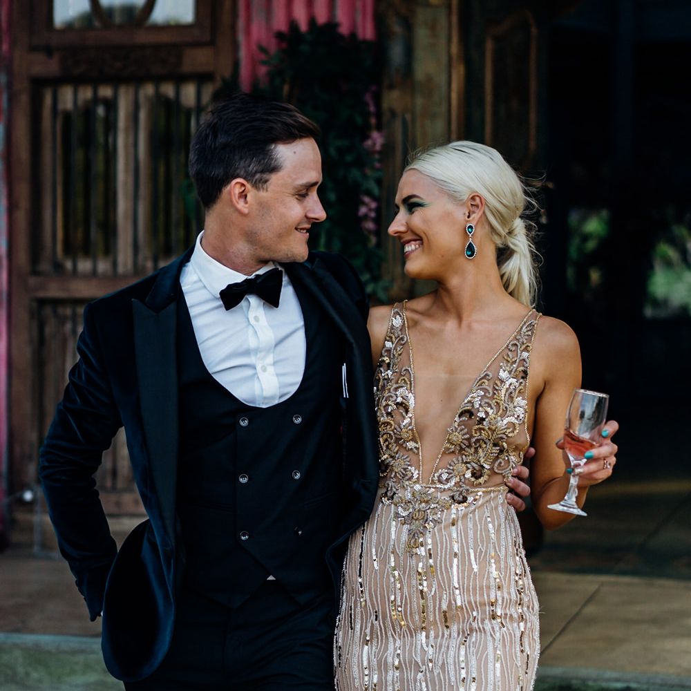 Groom in a black tuxedo with horseshoe waistcoat and bow tie and bride in in a glitzy gold wedding dress with plunging neckline 