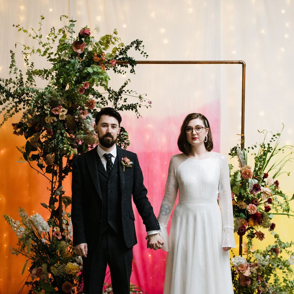 Bride in glasses and a sparkly Rolling In Roses wedding dress holding hands with her groom in wool suit in front of a copper pipe archway decorated with foliage and flowers in front of a tie dye fabric cloth