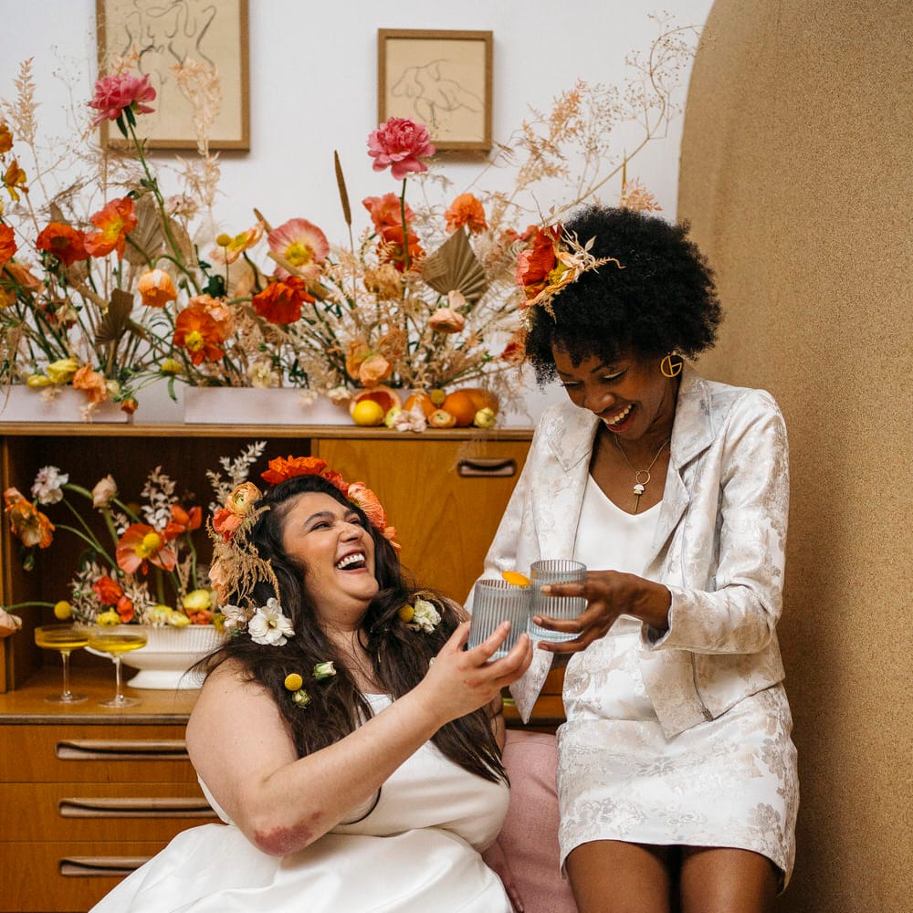 Two brides laughing in front of 70s wedding decor with floral display, both wearing bridal separates from Paper To Lace
