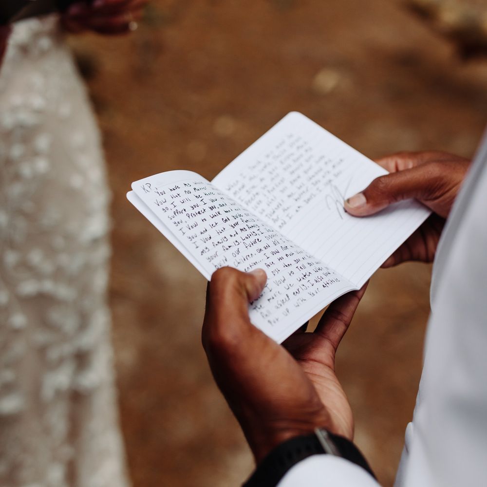 Groom holds notebook with private handwritten vows as he stands in woodland with bride in white embroidered Made With Love wedding dress at backyard wedding