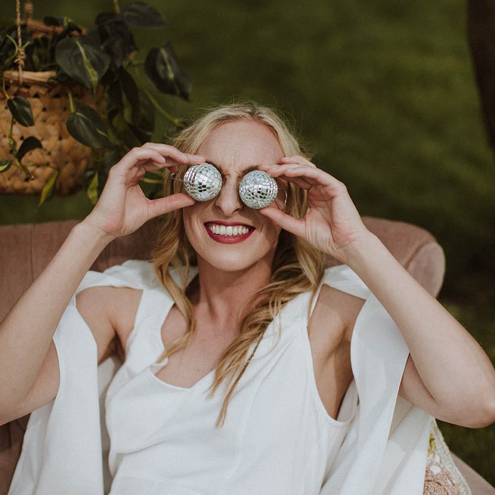 Bride in a jumpsuit and cape holding miniature disco balls at her eyes 