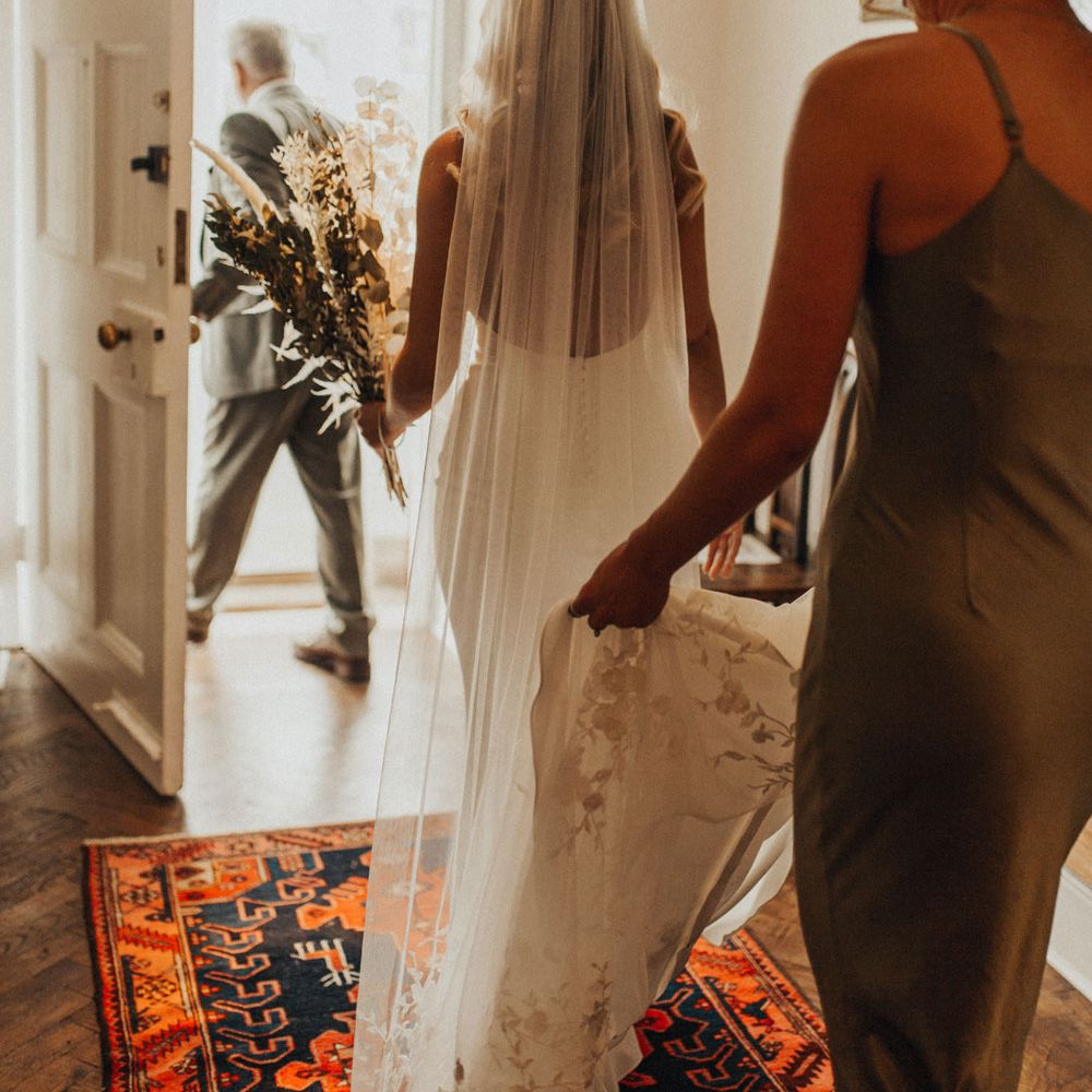 Bride in open back Made With Love wedding dress holding white and green dried bridal bouquet as she walks out of the front door as bridesmaid in satin sage green bridesmaids dress holds her veil before wedding at Anran Devon