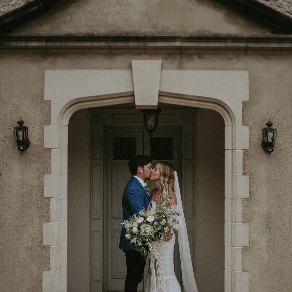 Bride in Grace Loves Lace bardot wedding dress and veil holding white and green bridal bouquet kisses groom in blue suit jacket in doorway at Isle of Wight wedding with macrame wedding decor