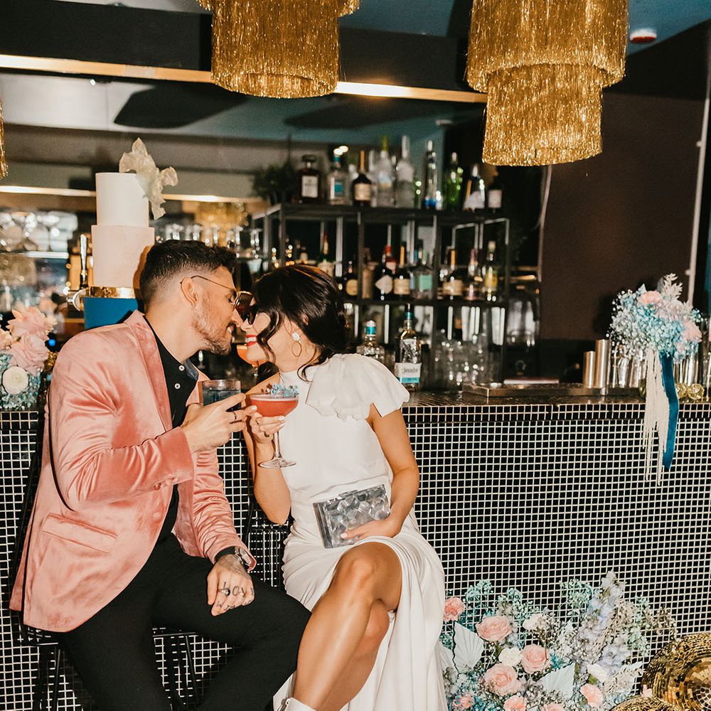 Stylish bride and groom sitting at the bar drinking cocktail in a pink velvet jacket and wedding dress with front split and boots 