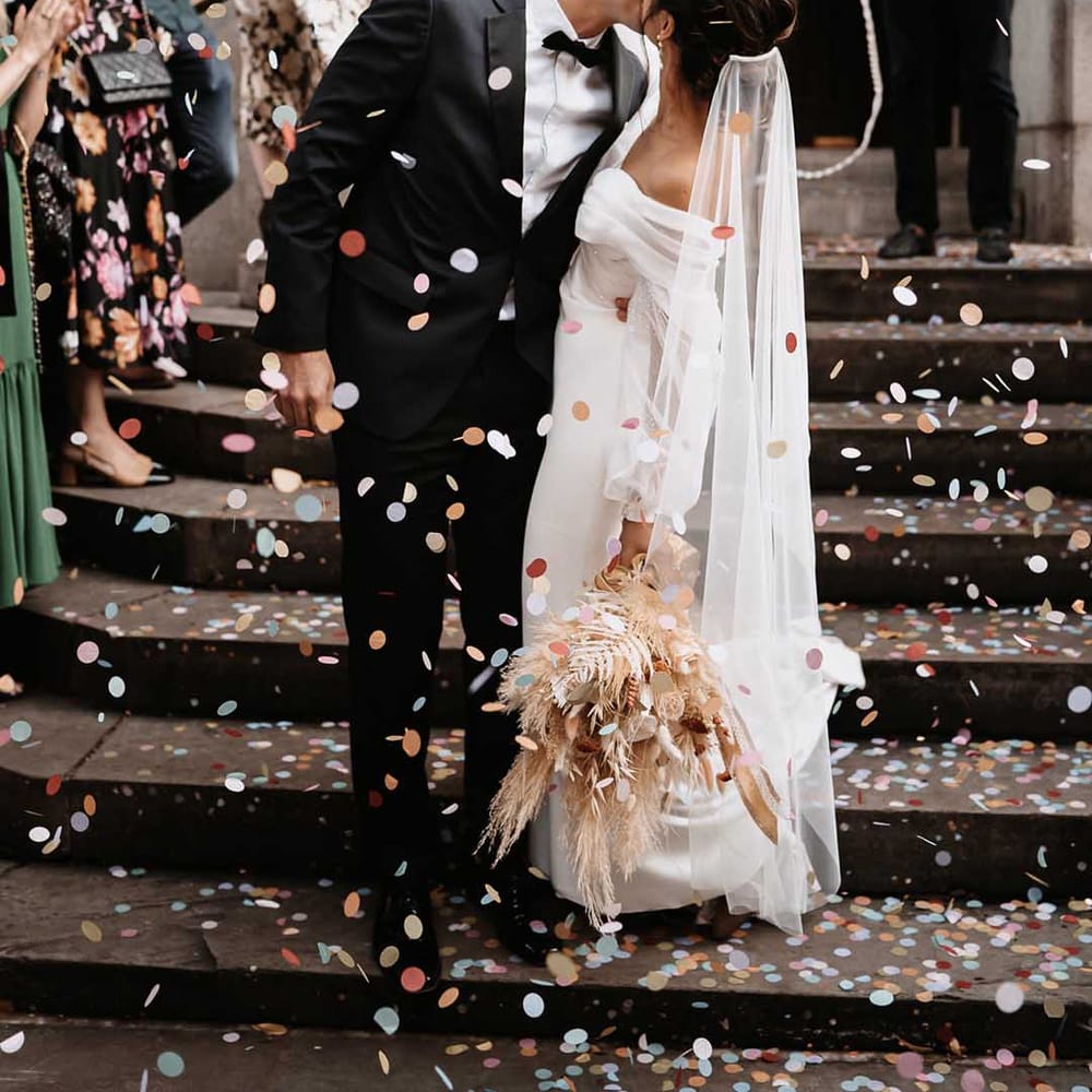 Bride and Groom celebrate with a kiss and pastel colourful confetti as their wedding guests clap with happiness 