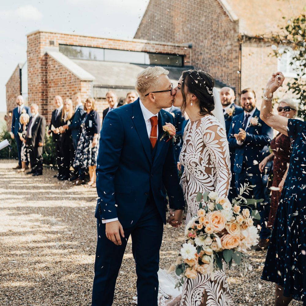 Bride with hair up do & Boho wedding dress kisses Groom in three piece navy suit with white shirt at Elmley Nature Reserve