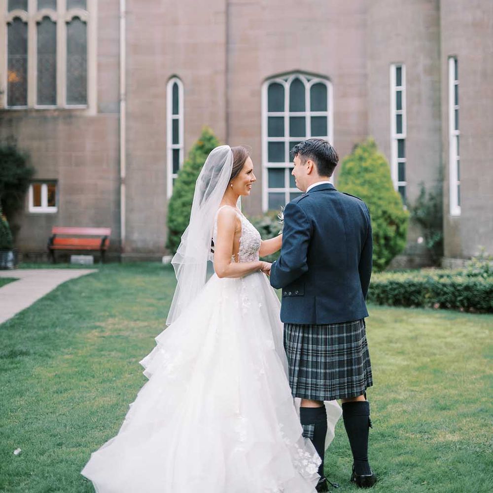 Bride in a layered tulle wedding dress and groom in a blue and green tartan kilt holding hands at Drumtochty castle 