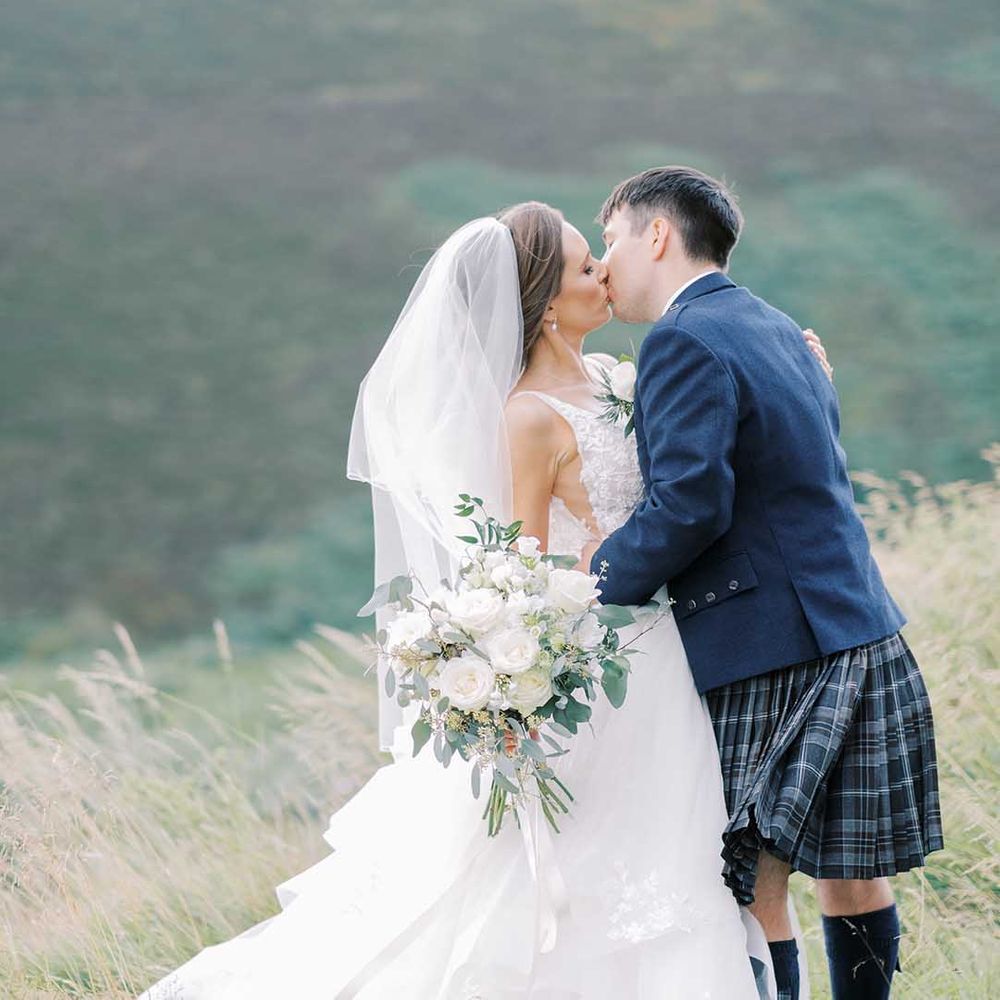 Groom in a tartan kilt and blue wool jacket kissing his bride in a princess wedding dress at their fairytale wedding 