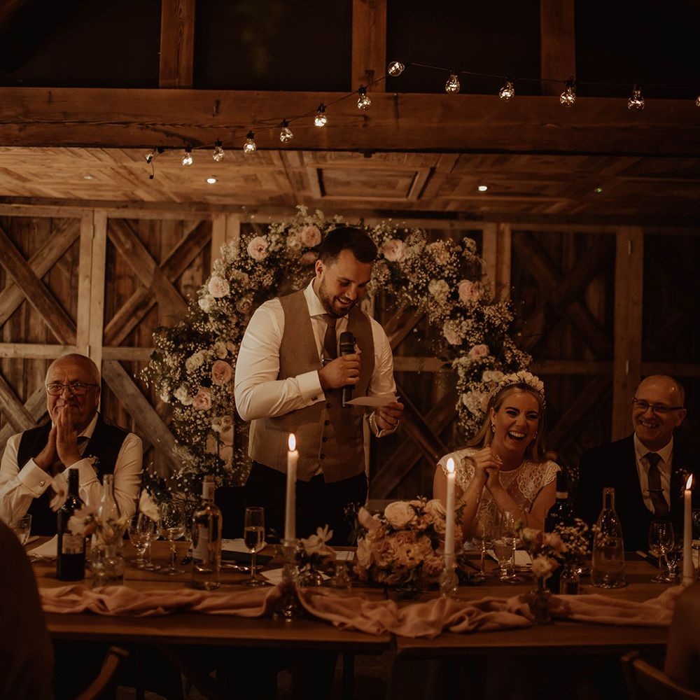 Groom in a waistcoat giving his wedding speech at Dewsall Court wedding with floral arch behind the top table 