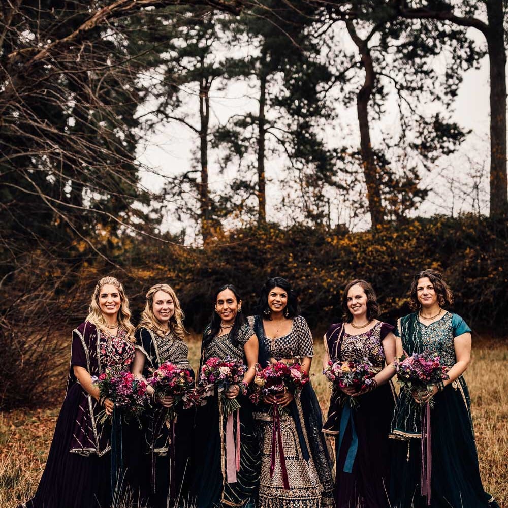 Bridesmaids in various Indian saree bridesmaid dresses all holding pink autumnal wedding bouquets with ribbon ties stand in field after wedding