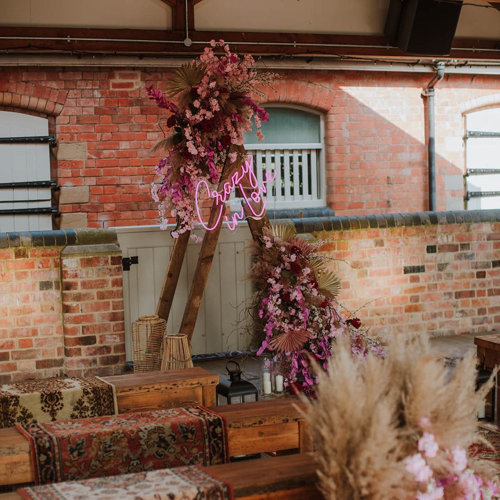 Prestwold Hall Barn wedding styling with wooden benches, wool rugs, lanterns and wooden frame altar decorated with a pink neon sign and flowers