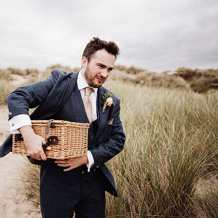 Bride in sheer long sleeve wedding dress carries her shoes along the beach as the groom carries wicker picnic basket