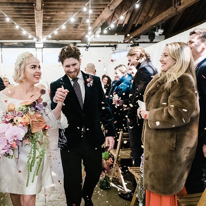 Bride in short wedding dress holding orchid wedding bouquet has confetti exit with groom in velvet suit jacket 
