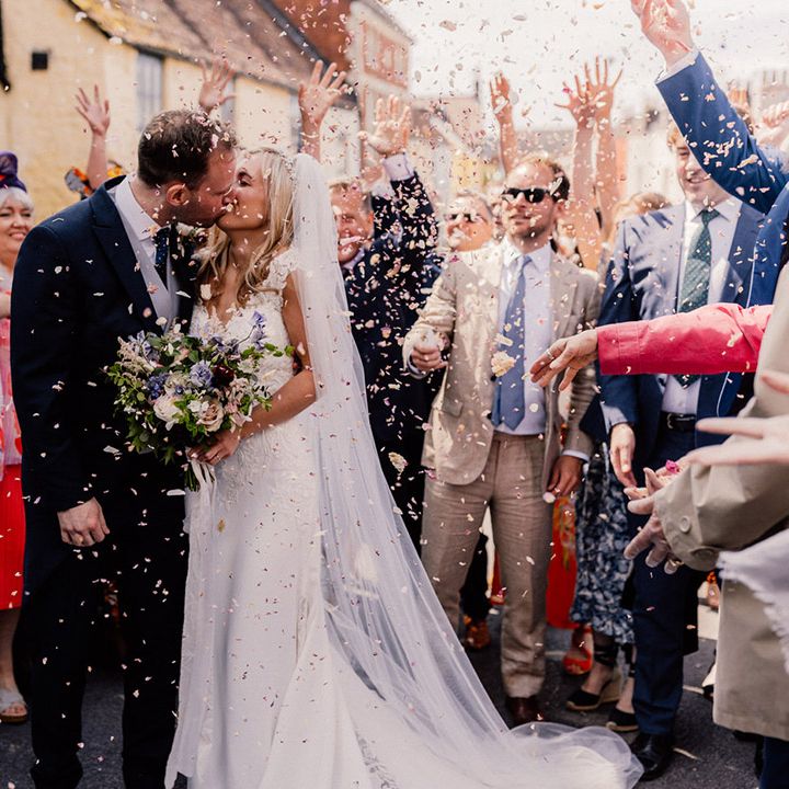 Bride and groom share a kiss outside their black and white wedding car as their guests throw confetti over them 