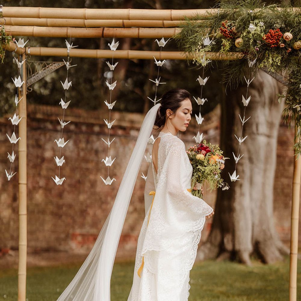 East Asian bride in a lace kimono style wedding dress and yellow dipped veil standing in front of a wooden bamboo frame decorated with paper cranes 