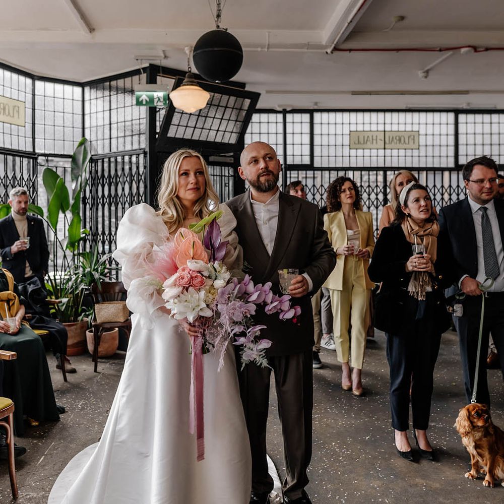 Emotional bride in a princess wedding dress with sheer sleeves holding a lilac orchid bouquet at her drinks reception at Floral Hall