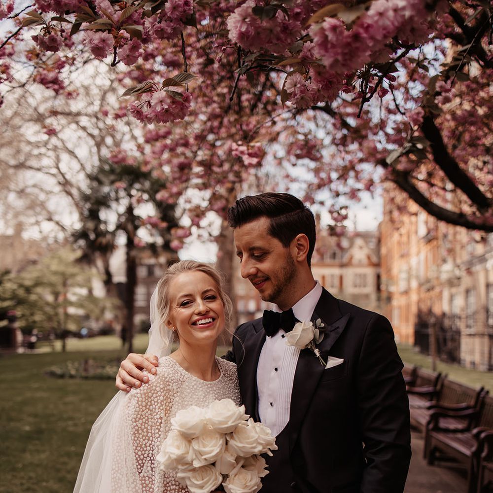 Bride in sparkly Jesus Peiro wedding dress and cape with groom in black tie unde spring blossom tree