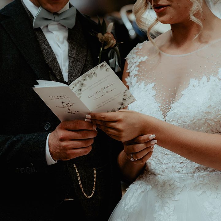 Bride in an illusion embellished wedding dress with the groom in a sage green bow tie reading through an order of service card