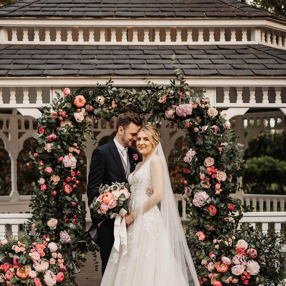 Groom in a dark suit embracing the bride in a lace wedding dress in front of the pink flower arch altar decor 