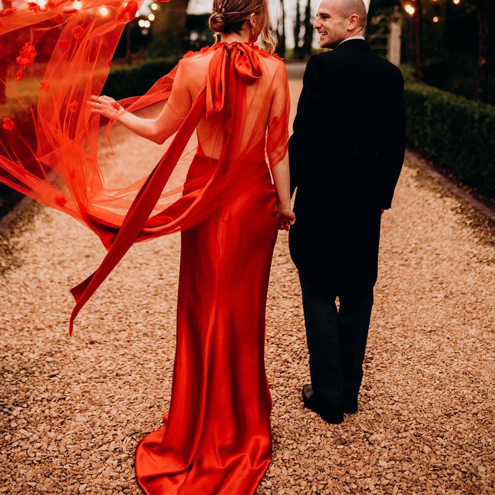 Bride in red satin wedding dress with cape and large red satin bow on the back neckline and groom in dark three piece suit walking hand in hand at Christmas wedding