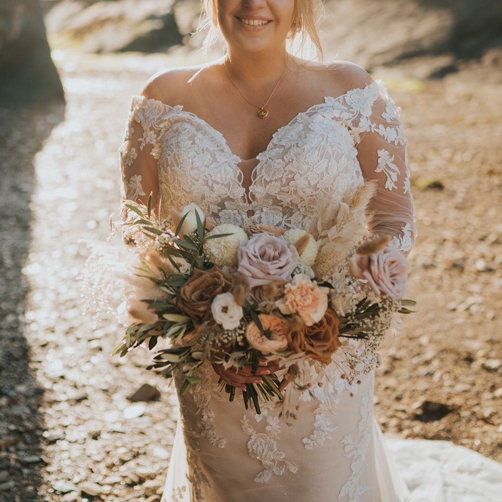 Bride wears off the shoulder long sleeve lace wedding dress whilst holding pastel bouquet 