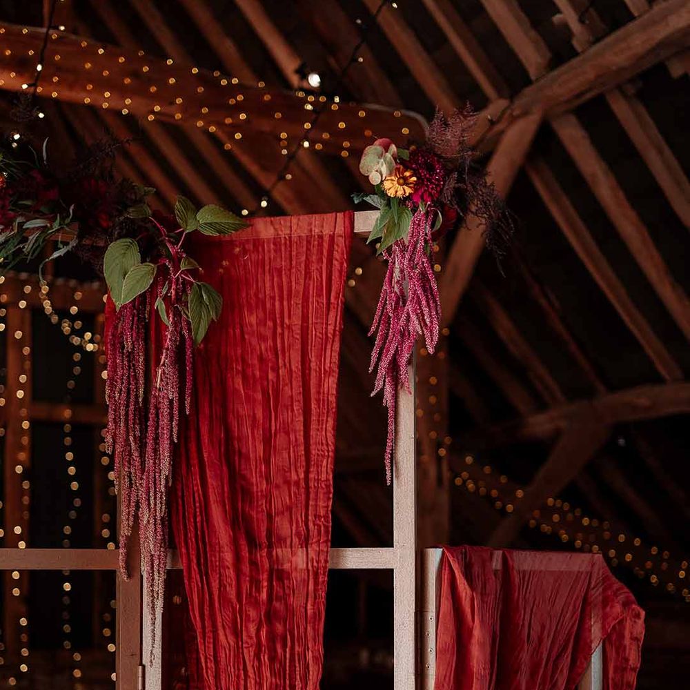 Deep red and burnt orange wedding drapes decor hung on ceiling beams with fairy lights and foliage and autumnal dried flower decorations