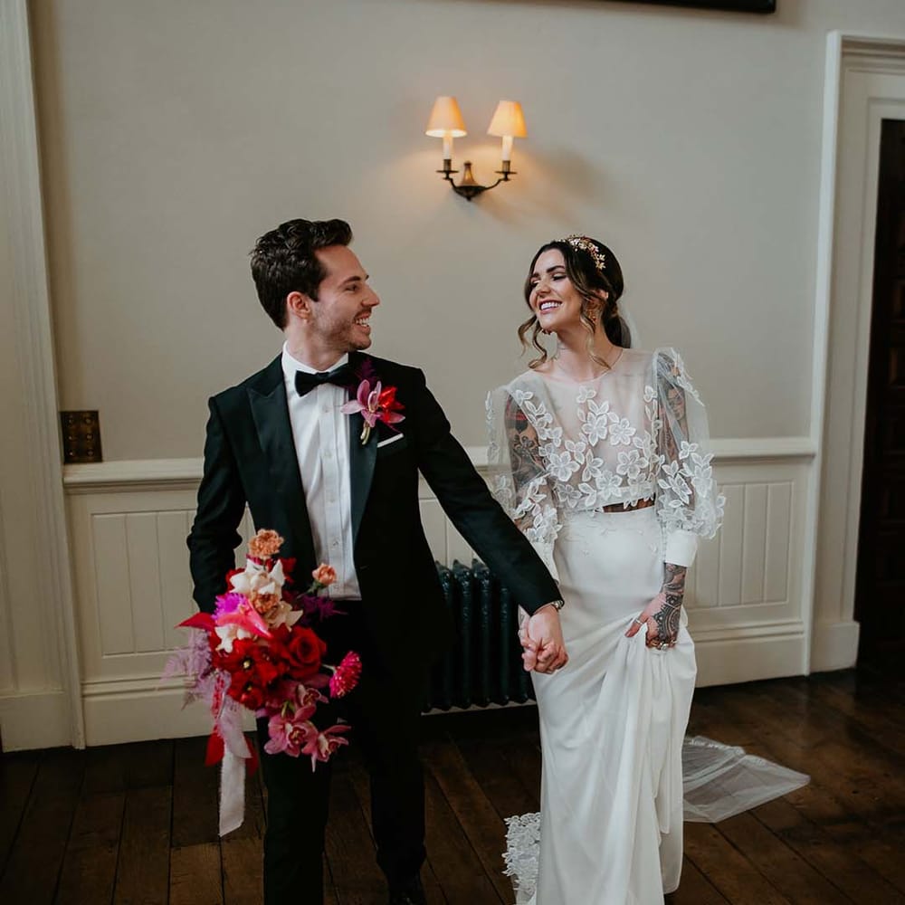 Bride in two piece bridal separates with puff sleeve sheer crop top with 3d appliqué flowers and full bodied silk skirt holding hands with groom wearing black tuxedo with magenta amaryllis and orchid boutonniere and holding hot pink and magenta floral bouquet