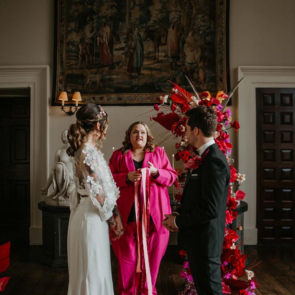 Groom in classic black tuxedo with magenta amaryllis and orchid boutonniere and bride in two piece bridal separates with puff sleeve sheer crop top with 3d appliqué flowers and full bodied silk skirt about to do handfasting ceremony with celebrant wearing hot pink suit