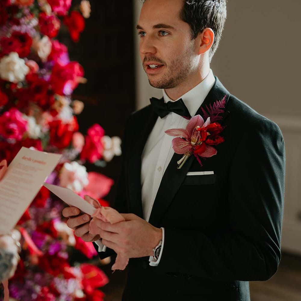 Groom reading his vows wearing classic black tuxedo with magenta amaryllis and orchid boutonniere 