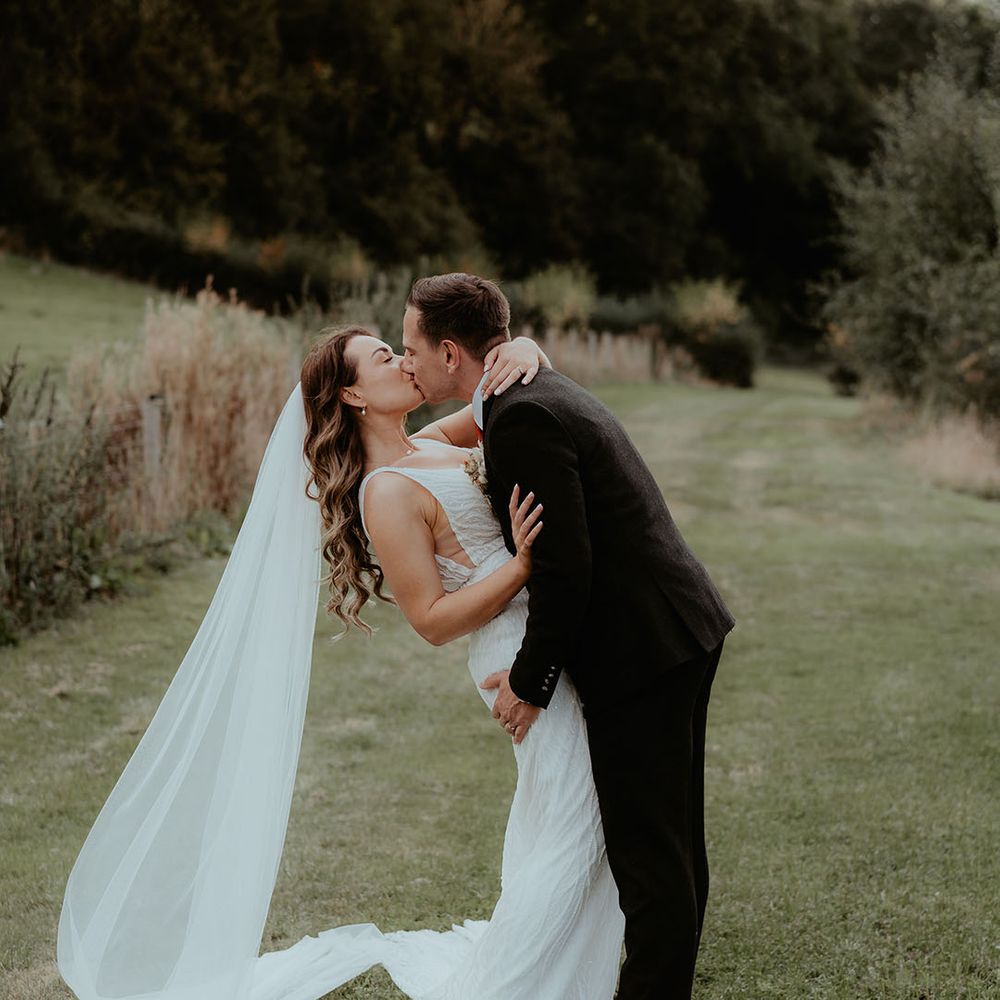 Groom in black suit kiss the bride as he dips her for cute couple portraits for the rustic glam wedding at Rhyse Farm 