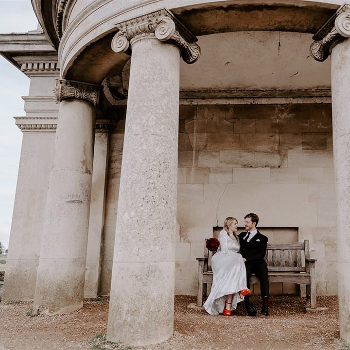 The bride in a metallic wedding dress wearing bright orange heels sitting on a bench with the groom in a pinstriped suit 