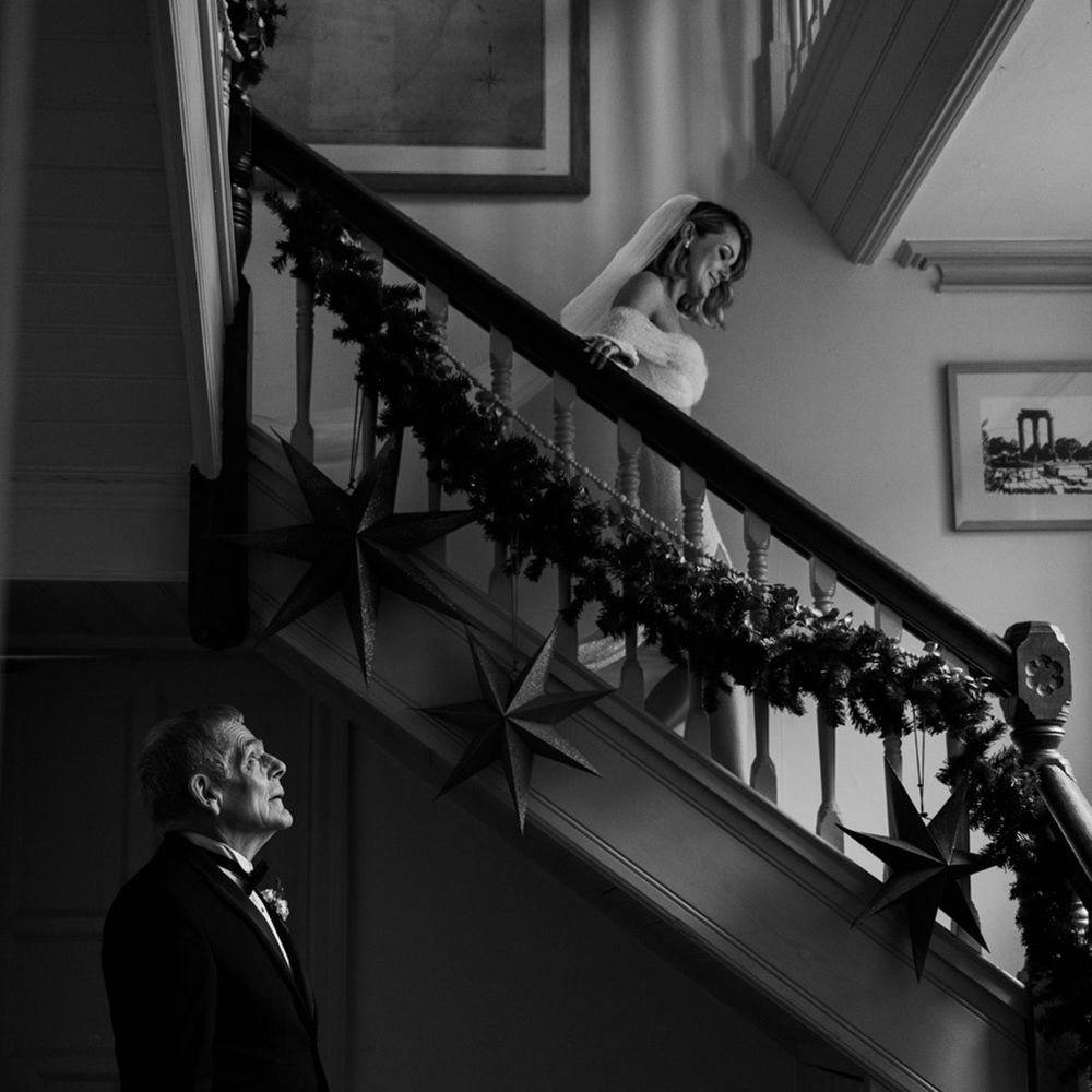 The bride begins walking down the stairs as the father of the bride in black tie waits to get his first look at her 