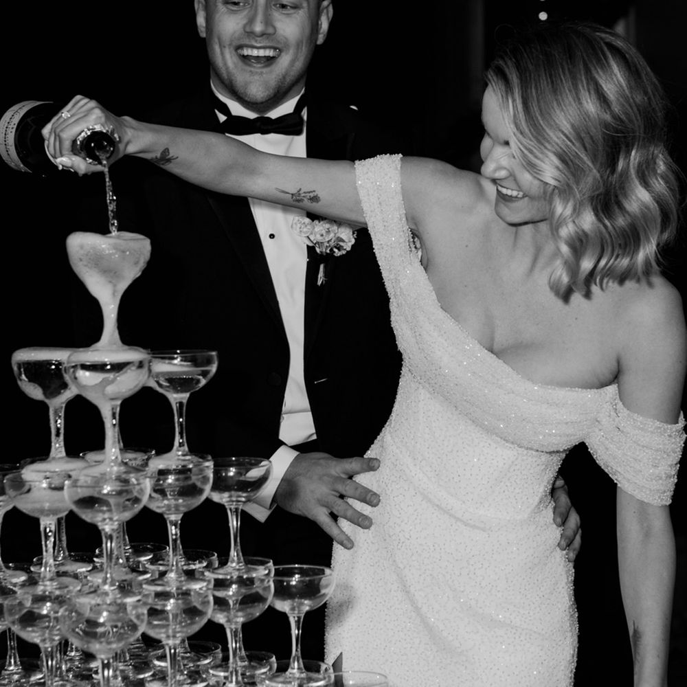 The groom embraces the bride with the bride in a white off the shoulder beaded wedding dress pouring champagne over the champagne tower 