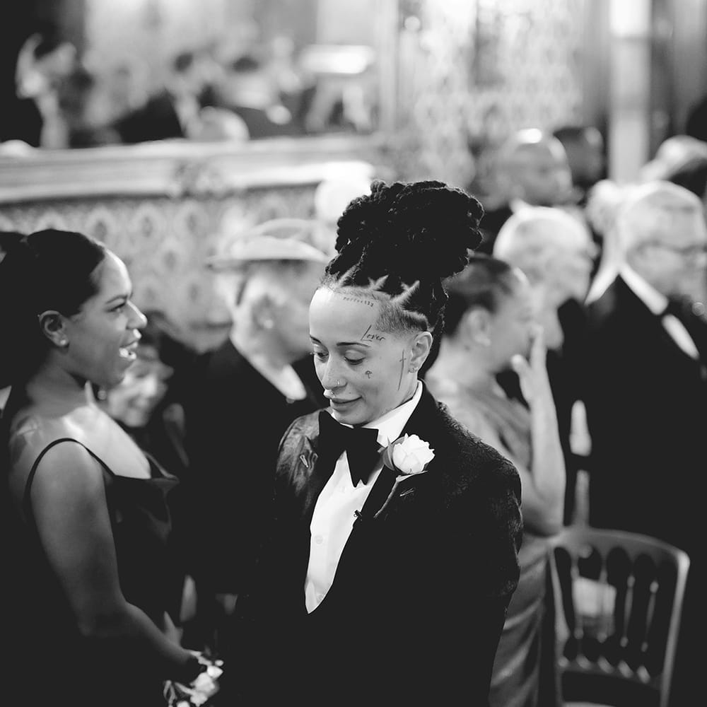 Bride in black tie tuxedo with black bowtie, white rose boutonniere waiting at the end of the aisle at Grittleton House