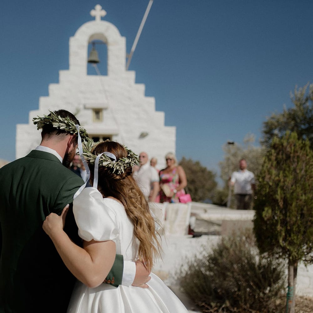 Bride and Groom embracing and wearing Stefana crowns in front of white greek chapel at destination wedding in Paros