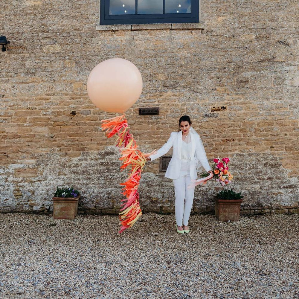 Bride in white bridal suit with closed toe bright green wedding heels holding large balloon with wedding streamers attached outside of Merriscourt wedding venue