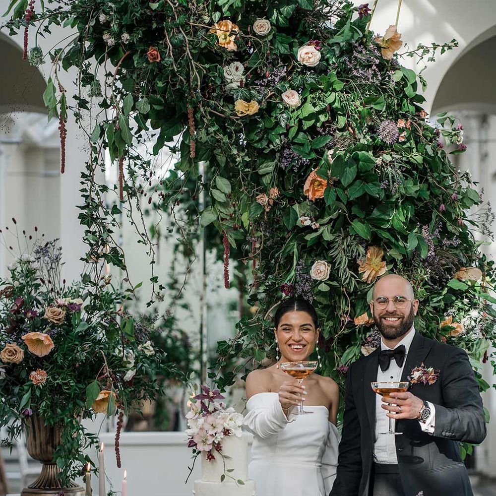 Bride in long sleeve off the shoulder wedding dress with pearl button detailing and a side slit standing with groom in classic black tuxedo with bowtie and wildflower boutonniere holding up cocktails standing by dessert table surrounded by suspended wildflower and foliage arrangements in large gold vases at Buxted Park