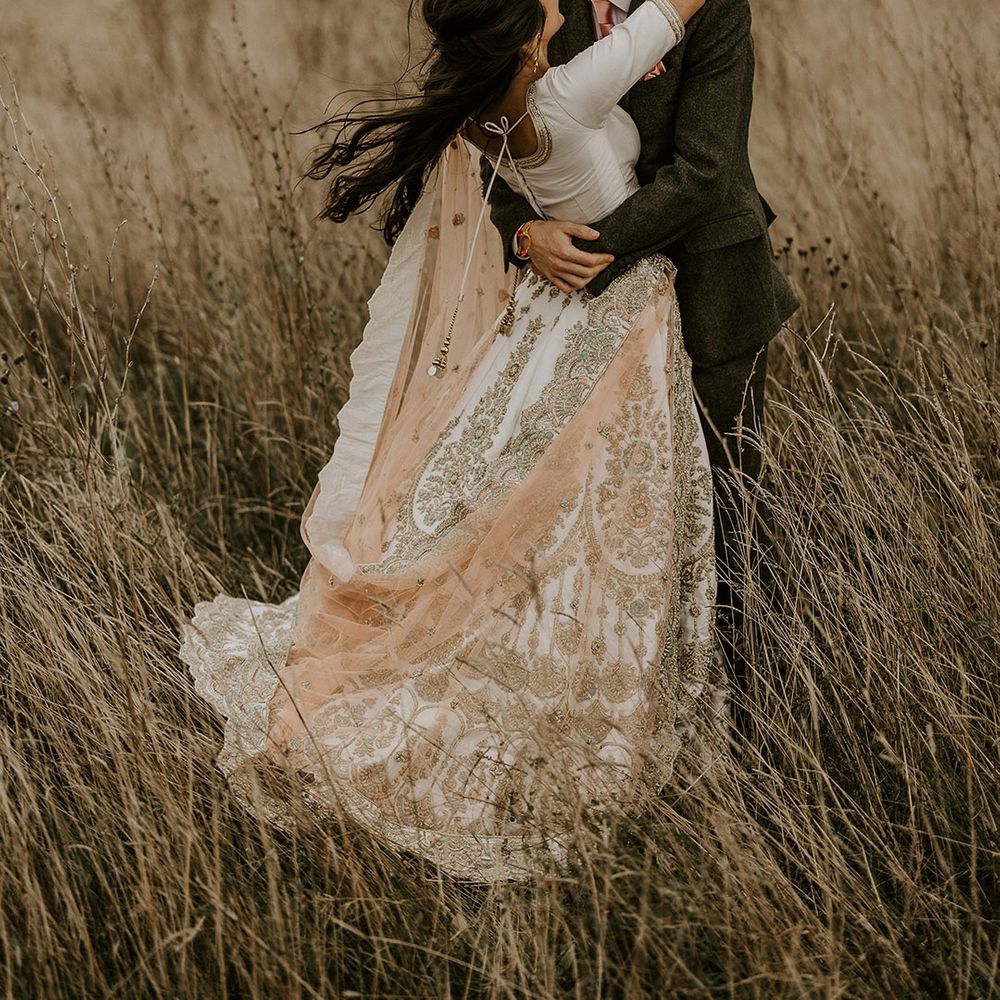 Bride in white and gold sparkly lehenga kissing groom in fields of Yorkshire countryside 