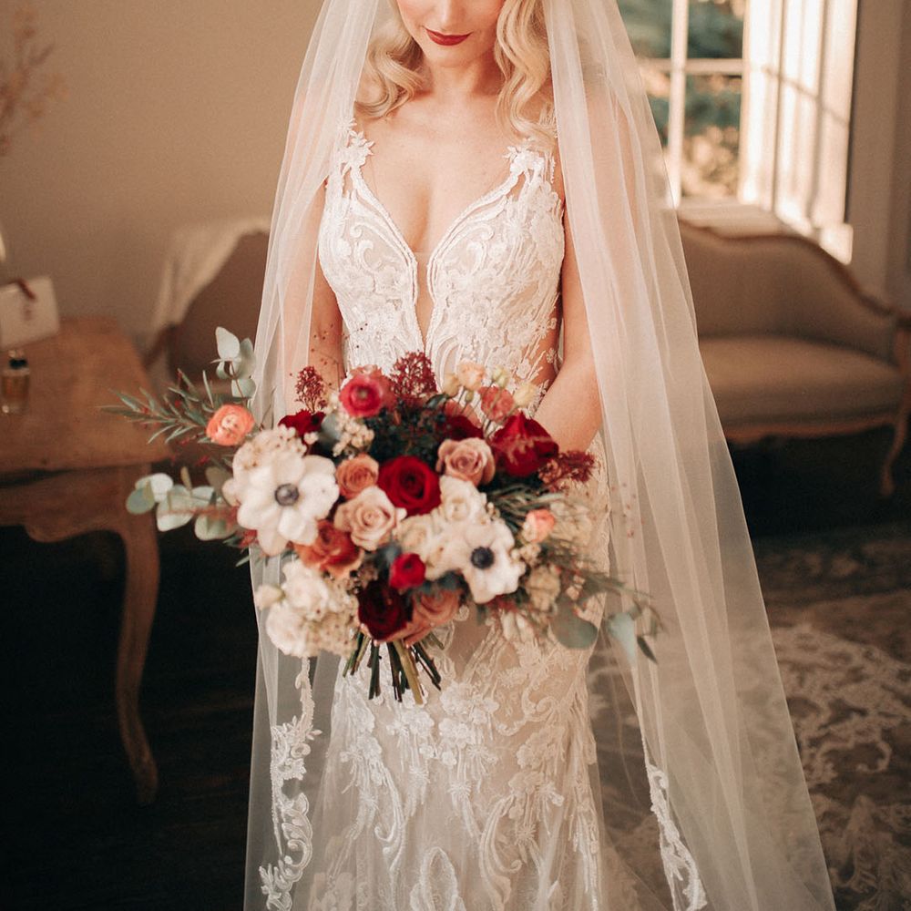 Bride wearing embellished lace wedding dress with embroidered veil holding white anemone and red rose bouquet 
