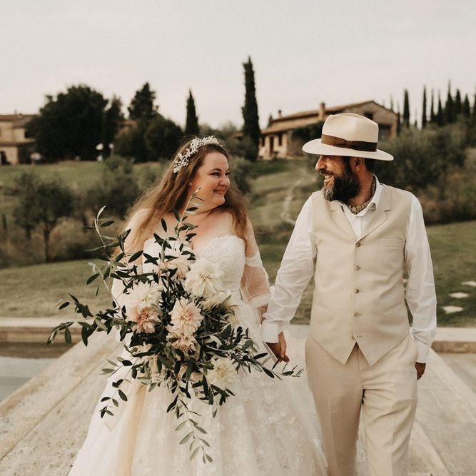 Bride and groom smiling, holding hands with an olive bouquet
