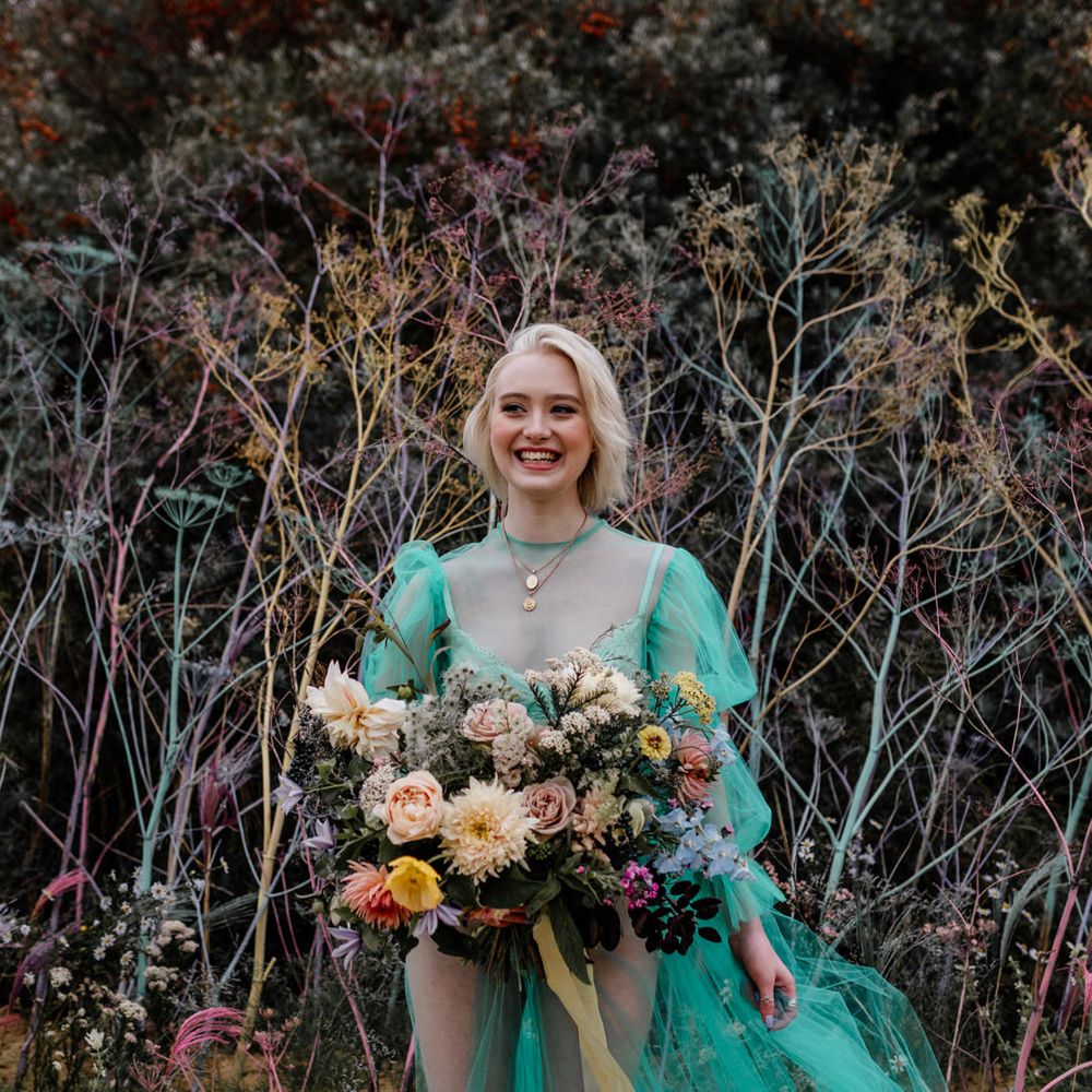 White bride with short blonde hair in a sheer pastel wedding dress on the beach holding an oversized colourful bouquet tied with ribbons