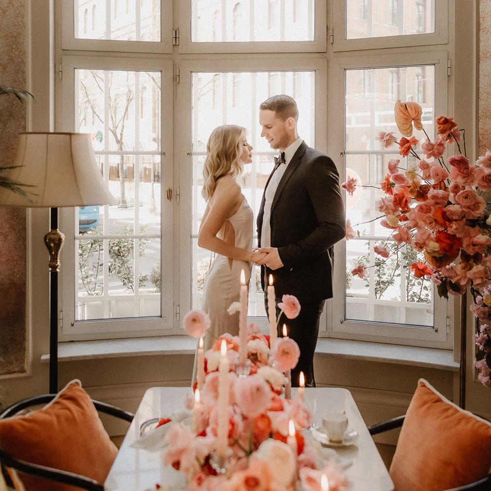 Bride and groom standing by an arched window, behind a table filled with pink and peach flowers